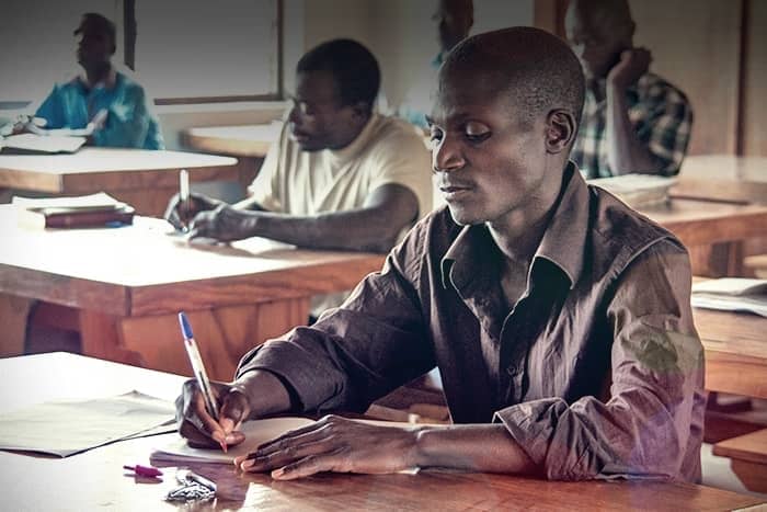 Ein Mann sitzt an einem hölzernen Schreibtisch und schreibt in ein Notizbuch, während andere an Tischen in einem Klassenzimmer arbeiten.