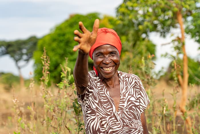 Lächelnde Frau mit rotem Kopftuch, die im Freien winkt, umgeben von Grünzeug und trockenen Pflanzen.