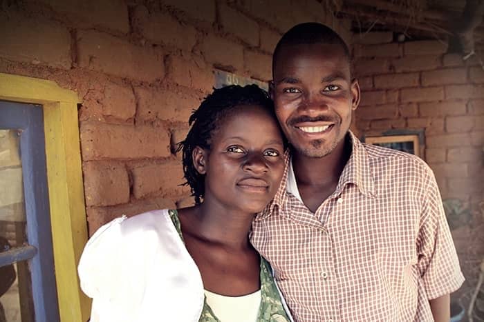 Ein lächelndes Paar steht dicht beieinander vor einer Backsteinmauer und einem Fenster mit gelb-blauem Rahmen.
