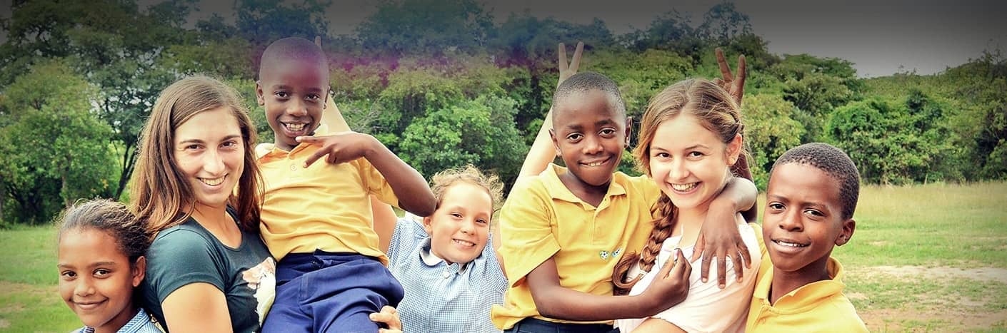 Eine Gruppe lächelnder Kinder und zwei Frauen posieren gemeinsam im Freien, einige mit Friedenszeichen, mit Grün im Hintergrund.