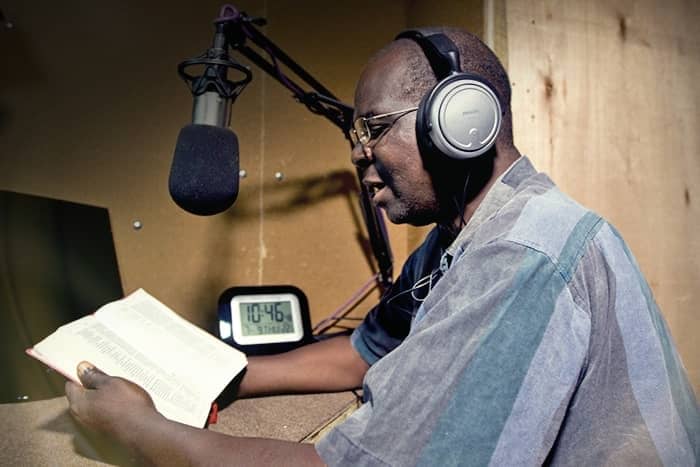 Ein Mann mit Kopfhörern liest in einem Aufnahmestudio aus einem Buch in ein Mikrofon. Eine Uhr zeigt 10:46.