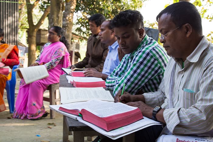 Die Menschen sitzen auf Bänken im Freien, lesen und schreiben in großen aufgeschlagenen Büchern, mit Bäumen und Sonnenlicht im Hintergrund.
