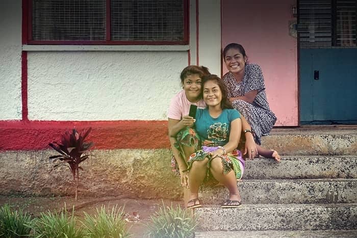 Drei Frauen, die lächelnd auf Betonstufen vor einem Gebäude mit roten und weißen Wänden sitzen.