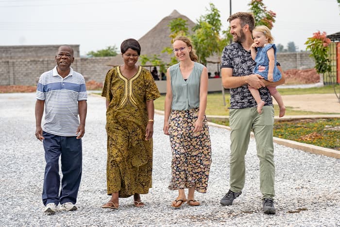 Vier Erwachsene und ein Kind spazieren und lächeln gemeinsam auf einem Kiesweg in der Nähe von Grünflächen und Gebäuden.