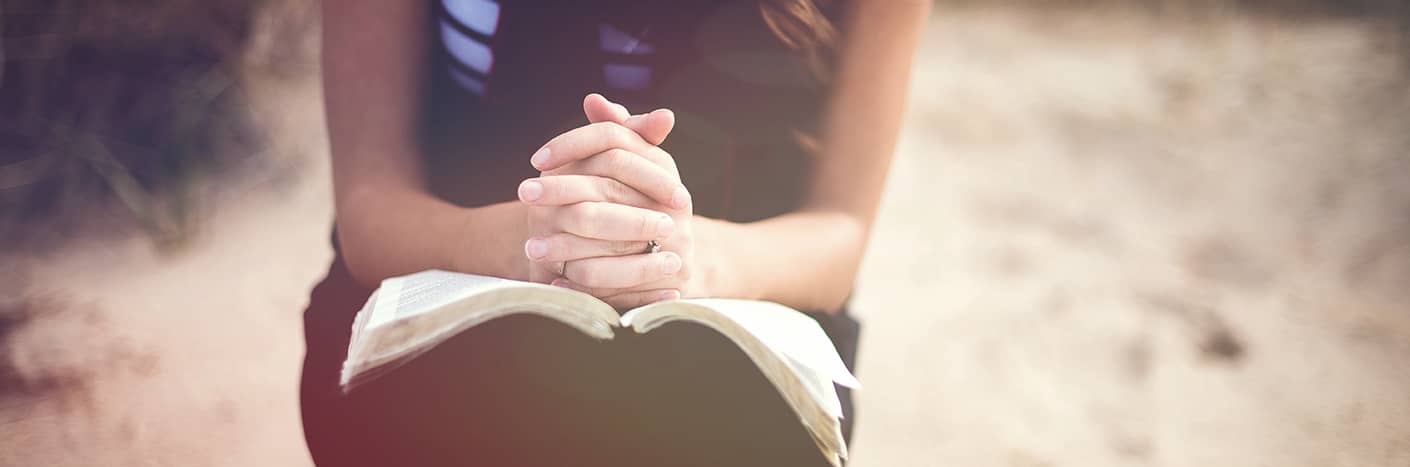 Nahaufnahme einer Person, die mit zum Gebet gefalteten Händen über einem aufgeschlagenen Buch, möglicherweise einer Bibel, im Freien sitzt.