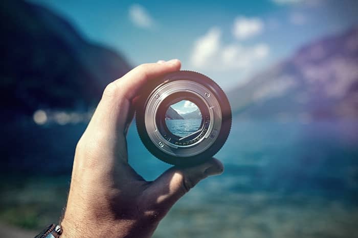 Eine Hand hält ein Kameraobjektiv, durch dessen Glas eine Berg- und Seeszene zu sehen ist.