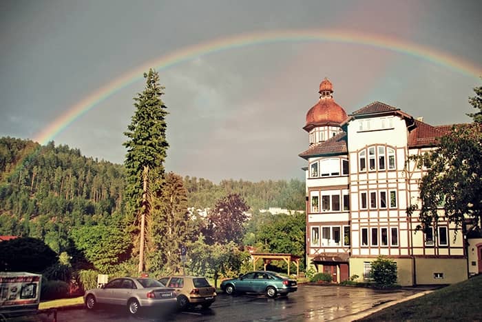 Ein Regenbogen wölbt sich an einem teilweise bewölkten Tag über ein historisches Gebäude, geparkte Autos und Bäume.