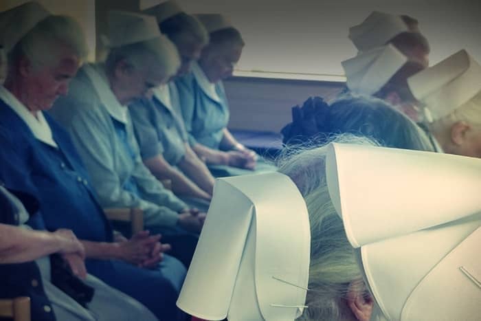 Ältere Frauen in Krankenschwesternkitteln und Hüten sitzen in Reihen, die Köpfe gesenkt, die Hände gefaltet, in einem schwach beleuchteten Raum.
