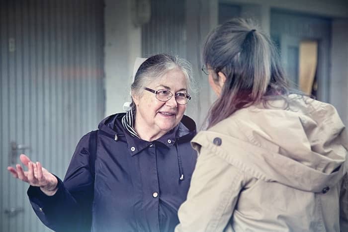 Zwei Frauen in Jacken unterhalten sich draußen, eine lächelt und gestikuliert mit der Hand, während die andere aufmerksam zuhört.