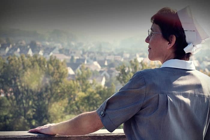 Eine Krankenschwester in Uniform steht im Freien und blickt nachdenklich auf eine Stadt mit Bäumen und Häusern im Hintergrund.