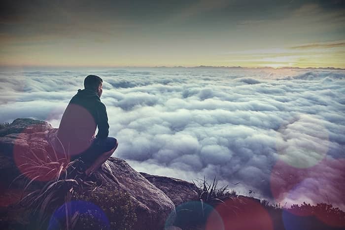 Eine Person sitzt auf einer Felsklippe und blickt bei Sonnenaufgang oder Sonnenuntergang über ein Wolkenmeer.