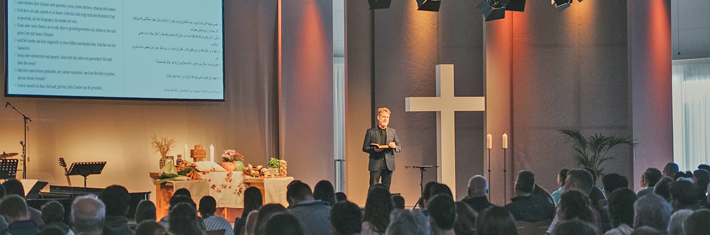 Eine Person spricht auf einer Bühne in einer Kirche mit einem großen Kreuz, Kerzen und projiziertem Text; das Publikum sitzt.