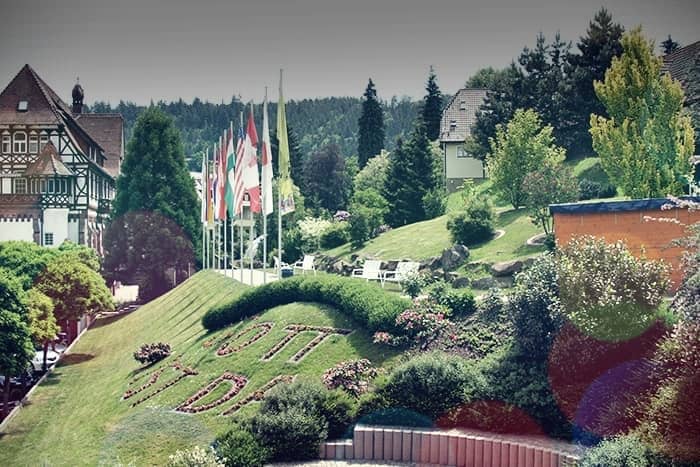 Eine Reihe von Fahnen und dekorativen Blumen auf einem Hügel verkünden "OTTO" in einem Landschaftsgarten in der Nähe traditioneller europäischer Gebäude.