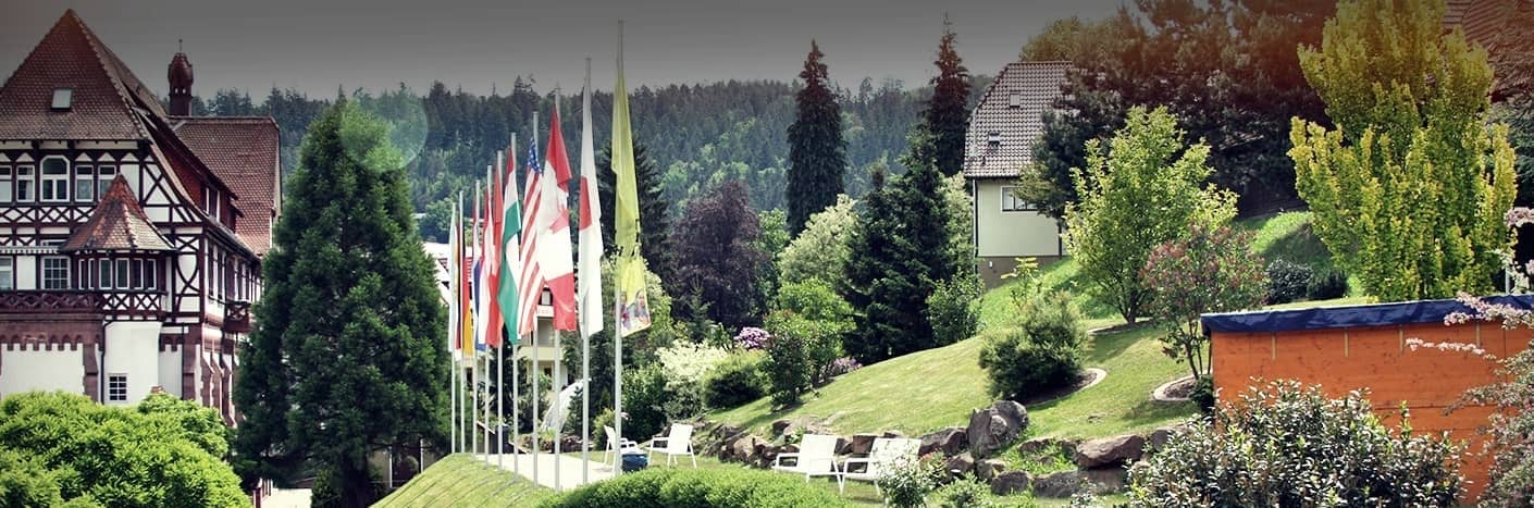 Eine Reihe internationaler Flaggen steht zwischen Bäumen und Gebäuden an einem üppigen, grünen Hügel in malerischer Umgebung.