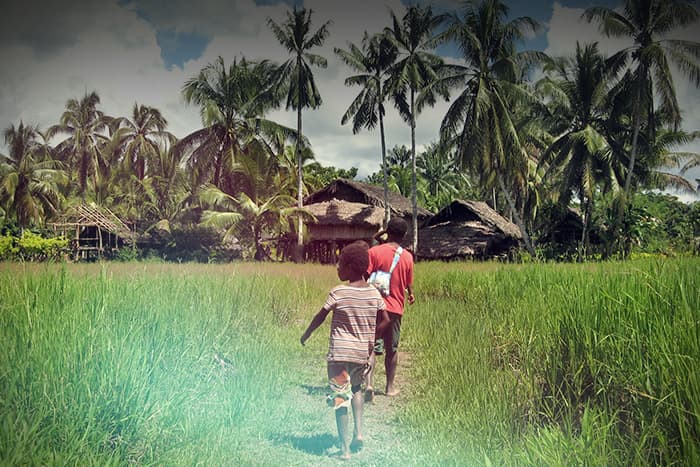 Zwei Personen gehen auf einem grasbewachsenen Weg zu strohgedeckten, von Palmen umgebenen Hütten in einer ländlichen, tropischen Umgebung.