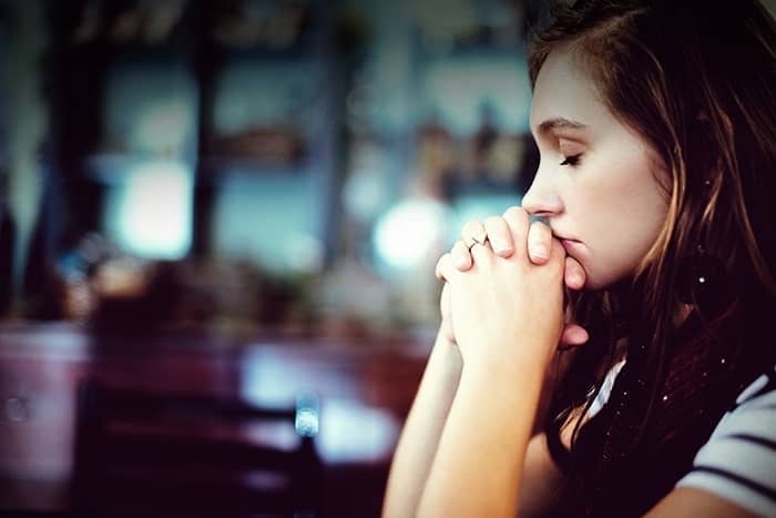 Junge Frau, die mit geschlossenen Augen und gefalteten Händen sitzt und in Gedanken oder im Gebet versunken zu sein scheint.