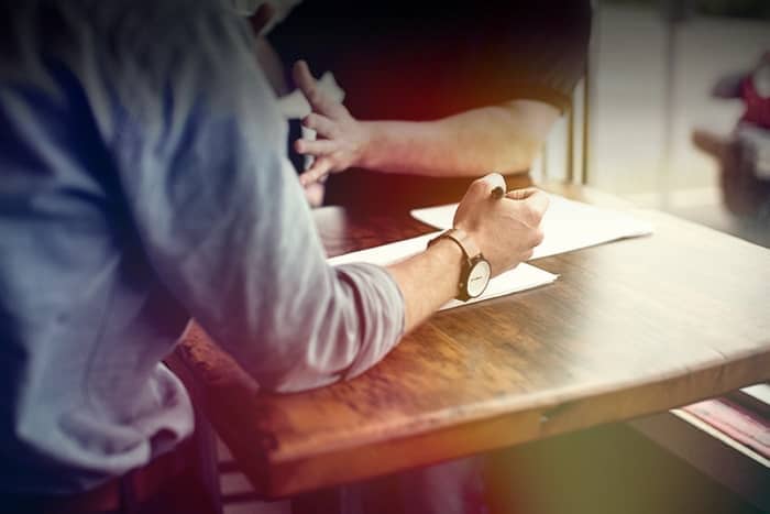 Zwei Menschen sitzen an einem Holztisch, einer schreibt auf einem Papier, und unterhalten sich an einem sonnendurchfluteten Fenster.