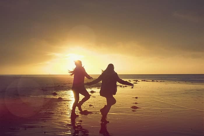Zwei Menschen, die sich an den Händen halten und bei Sonnenuntergang über einen Strand laufen, der sich vom hellen Himmel abhebt.