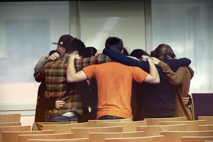 Eine Gruppe von Personen steht in einem Klassenzimmer in einem Kreis und legt die Arme umeinander.