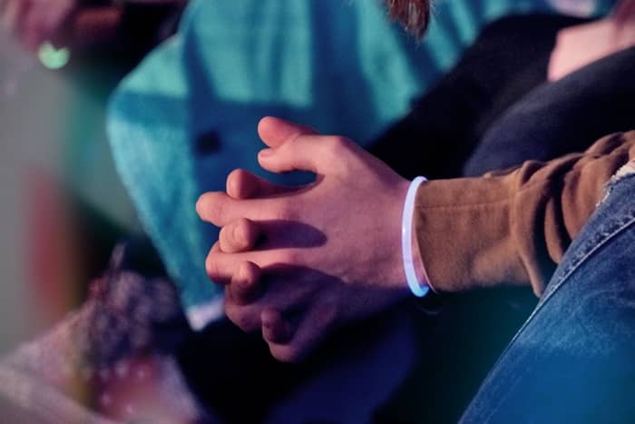 Nahaufnahme von zwei sich an den Händen haltenden Personen, von denen eine einen braunen Ärmel und ein leuchtendes Armband trägt, vor blauem Hintergrund.