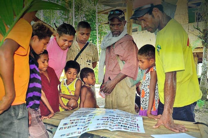 Eine Gruppe von Menschen sitzt um einen Tisch im Freien und sieht sich Papiere mit Fotos und Texten an.