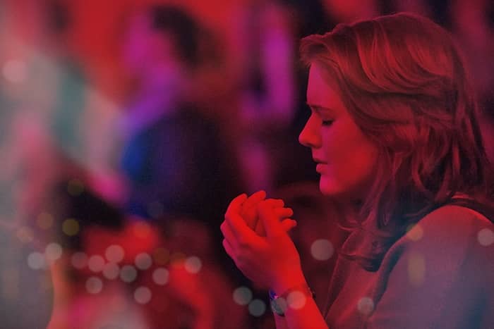 Eine Frau mit geschlossenen Augen hält ihre Hände zum Gebet zusammen, umgeben von weichen, bunten, unscharfen Lichtern.