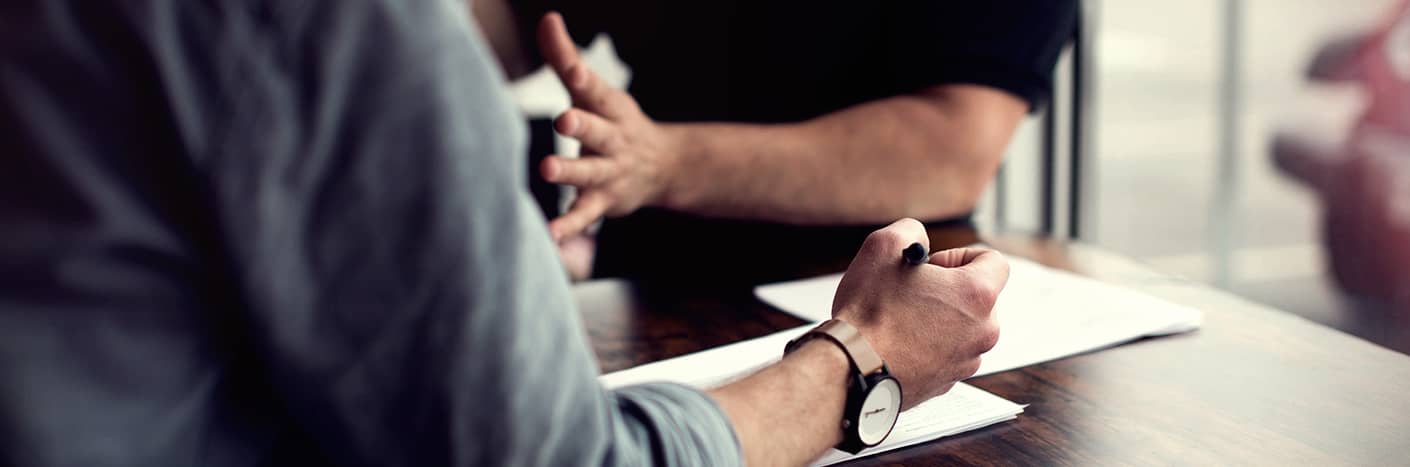 Zwei Personen sitzen mit Papieren an einem Tisch, einer schreibt, der andere gestikuliert während einer Diskussion.