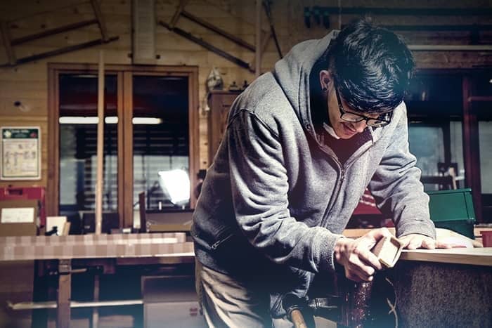 Eine Person mit Brille schleift Holz in einer Werkstatt, umgeben von Holzbearbeitungswerkzeugen und -geräten.