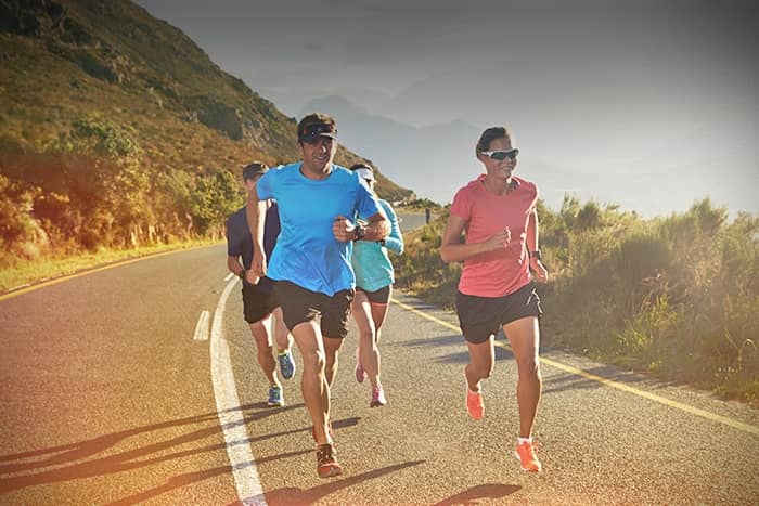 Vier Personen laufen auf einer kurvigen Straße im Freien, tragen Sportkleidung und Sonnenbrillen, mit Hügeln und Sonnenlicht im Hintergrund.