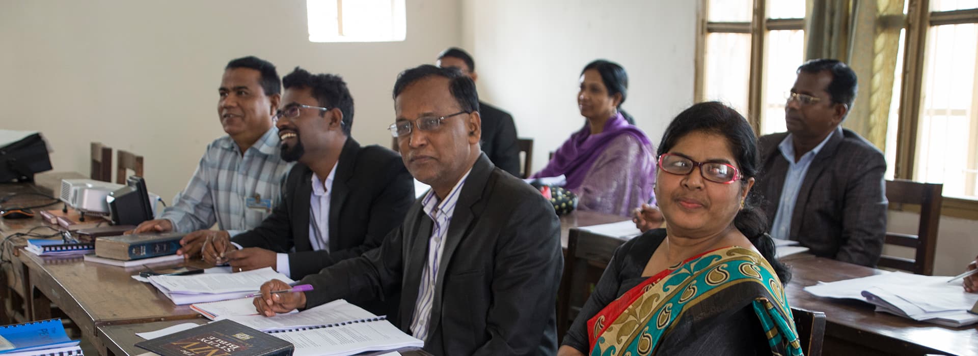 Menschen, die an Tischen in einem Klassenzimmer sitzen, einige lächelnd, mit Heften und Büchern vor sich.