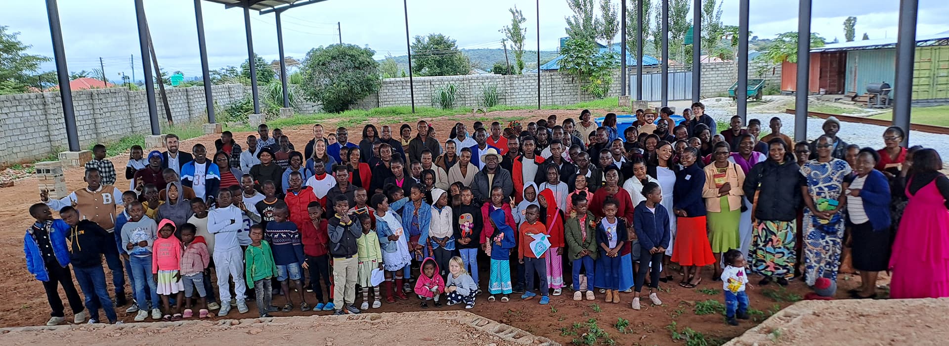 Eine große Gruppe von Menschen, darunter Kinder und Erwachsene, posiert im Freien unter einem metallüberdachten Unterstand.