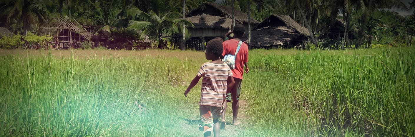 Zwei Kinder gehen auf einem Pfad durch hohes Gras auf strohgedeckte Häuser in einem ländlichen Dorf zu.