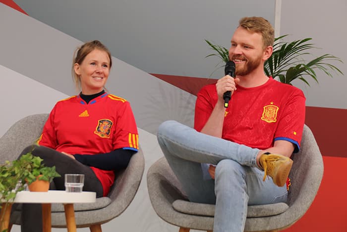 Zwei Personen in roten spanischen Fußballtrikots sitzen auf der Bühne und unterhalten sich; der Mann hält ein Mikrofon und lächelt.