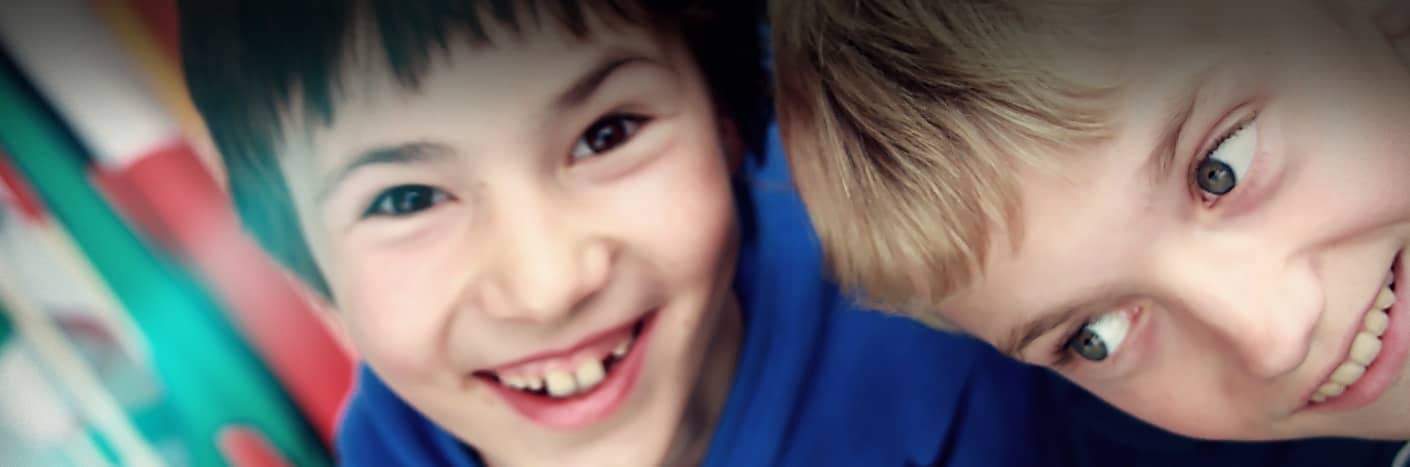 Zwei Jungen lächeln und lachen eng beieinander, beide tragen blaue Oberteile, mit einem bunten Hintergrund.
