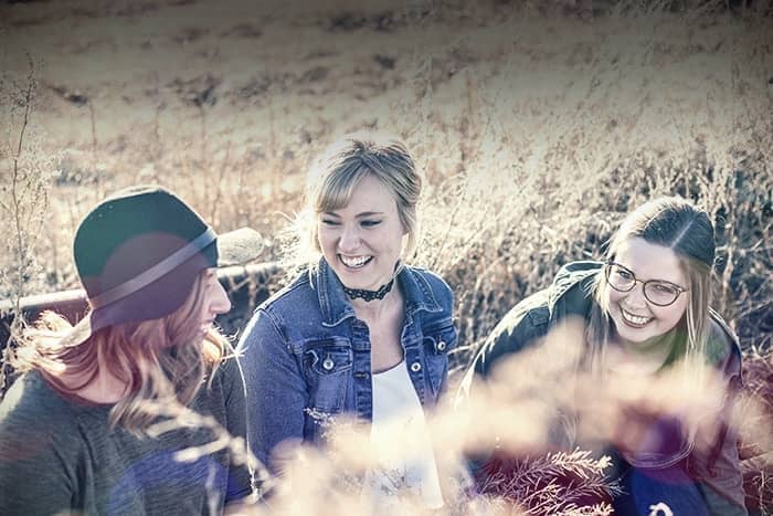 Drei junge Frauen sitzen an einem sonnigen Tag draußen auf einer Wiese und lächeln und lachen zusammen.