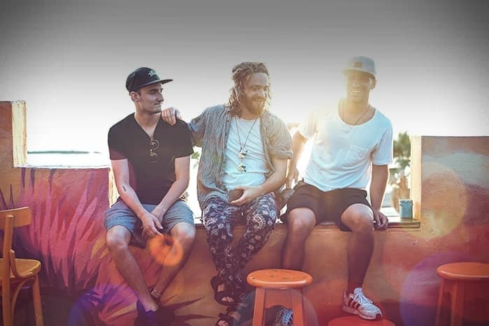 Drei Männer sitzen zusammen und lächeln auf einer bunten Außenwand mit orangefarbenen Stühlen und einem künstlerischen Wandgemälde im Hintergrund.