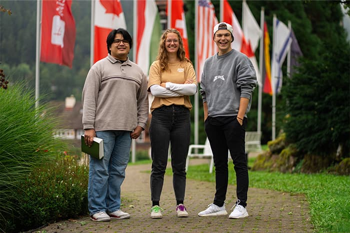 Drei lächelnde junge Erwachsene stehen draußen auf einem Weg mit verschiedenen internationalen Flaggen im Hintergrund.