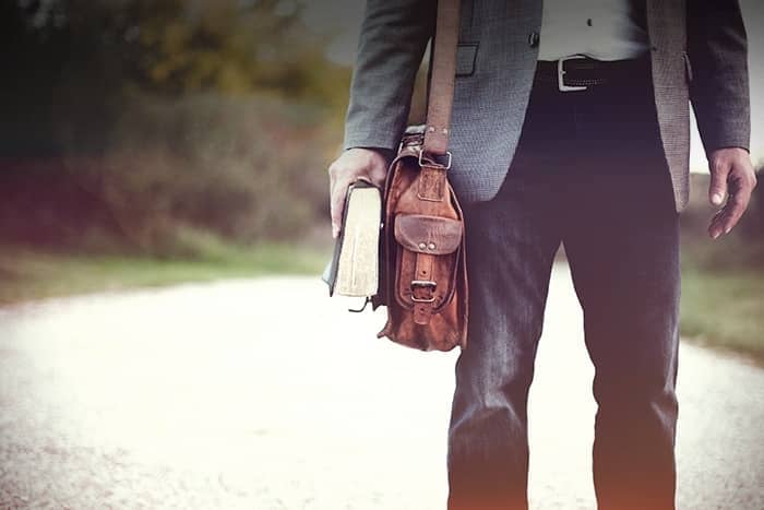 Mann steht im Freien mit einem Buch und einer braunen Ledertasche in der Hand, bekleidet mit Jeans und einem grauen Blazer.