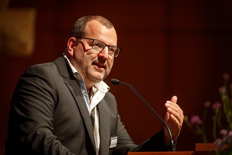 Ein Mann im dunklen Anzug und mit Brille spricht an einem Podium mit Mikrofon und gestikuliert mit der Hand.