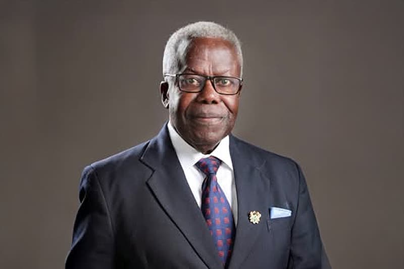 Ein älterer Mann in einem dunklen Anzug, mit Brille und Krawatte steht vor einem schlichten Hintergrund und lächelt leicht.