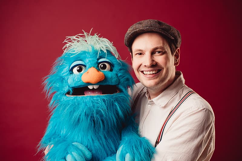 Ein lächelnder Mann mit einer flachen Mütze hält eine blaue, pelzige Marionette mit großen Augen und einer orangefarbenen Nase vor einem roten Hintergrund.