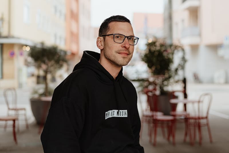 Ein Mann mit Brille und schwarzem Kapuzenpulli sitzt draußen in einem Café mit roten Stühlen und Tischen und schaut in die Kamera.