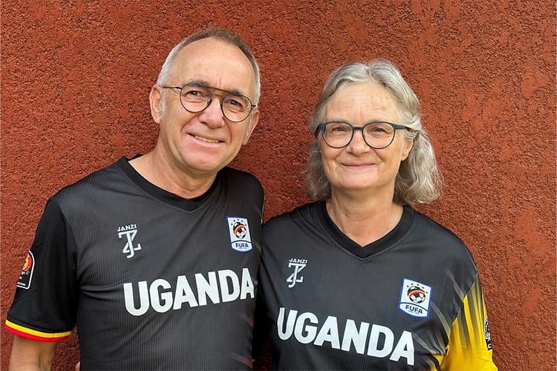 Zwei lächelnde Menschen in schwarzen Uganda-Football-Trikots stehen nebeneinander vor einer rot strukturierten Wand.