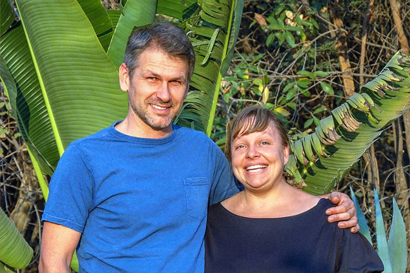 Ein lächelnder Mann und eine lächelnde Frau stehen zusammen im Freien mit großen grünen tropischen Blättern im Hintergrund.