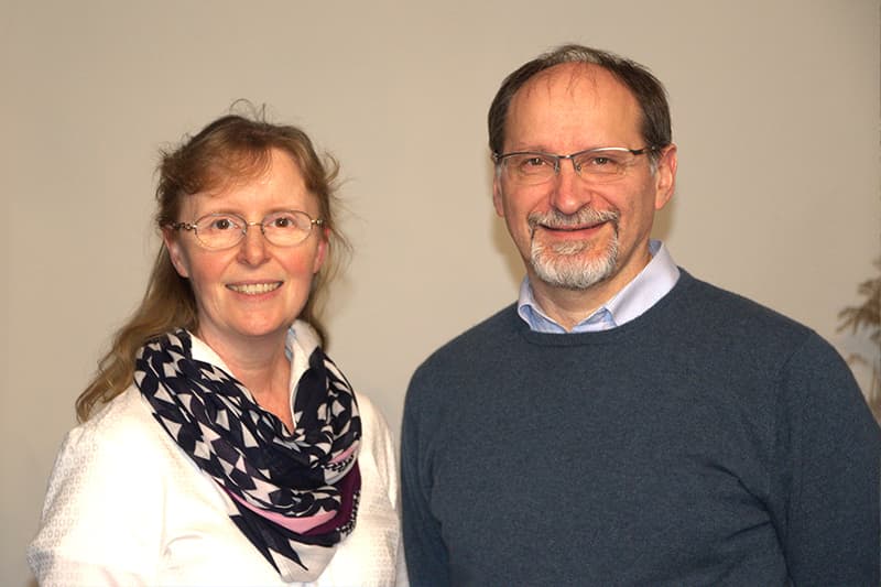 Eine Frau und ein Mann stehen lächelnd nebeneinander vor einem einfarbig beigen Hintergrund.
