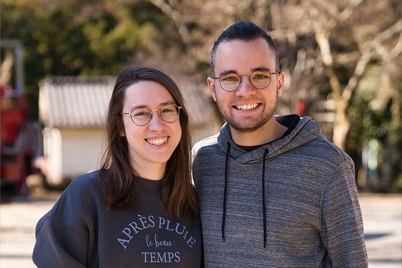 Zwei Personen mit Brille, die im Freien lächeln, stehen dicht beieinander, mit Bäumen und einem Haus im Hintergrund.