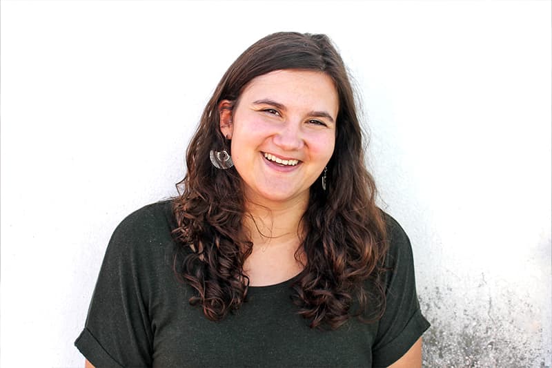 Frau mit langen braunen lockigen Haaren und dunklem Hemd, lächelnd vor einer weißen Wand.