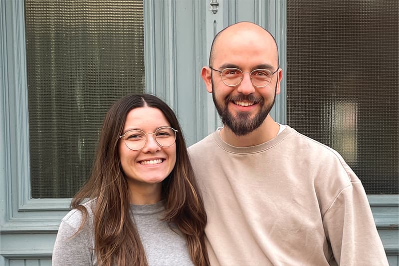 Zwei Personen mit Brillen, die lächelnd und dicht beieinander vor einer hellblauen Tür und einem Fenster stehen.