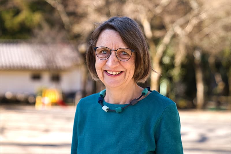 Lächelnde Frau mit Brille und blaugrünem Oberteil, die im Freien steht, mit Bäumen und einem Gebäude im Hintergrund.