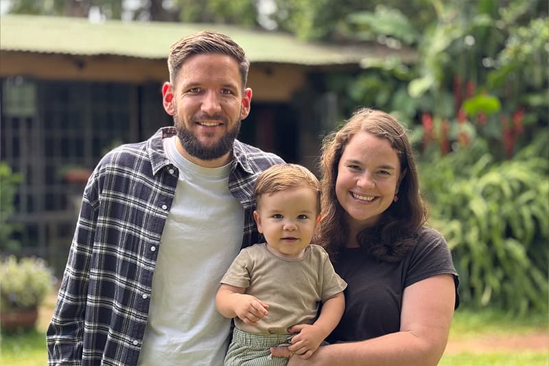 Ein lächelnder Mann und eine lächelnde Frau halten ein kleines Kind im Freien, mit Grünflächen und einem Gebäude im Hintergrund.